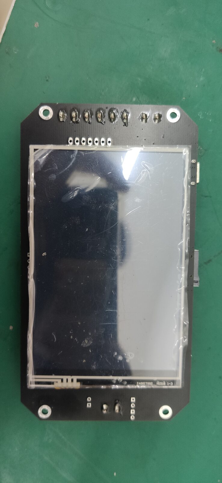 Small circuit board with a black glass touchscreen-style display in a black frame, on a green mat.