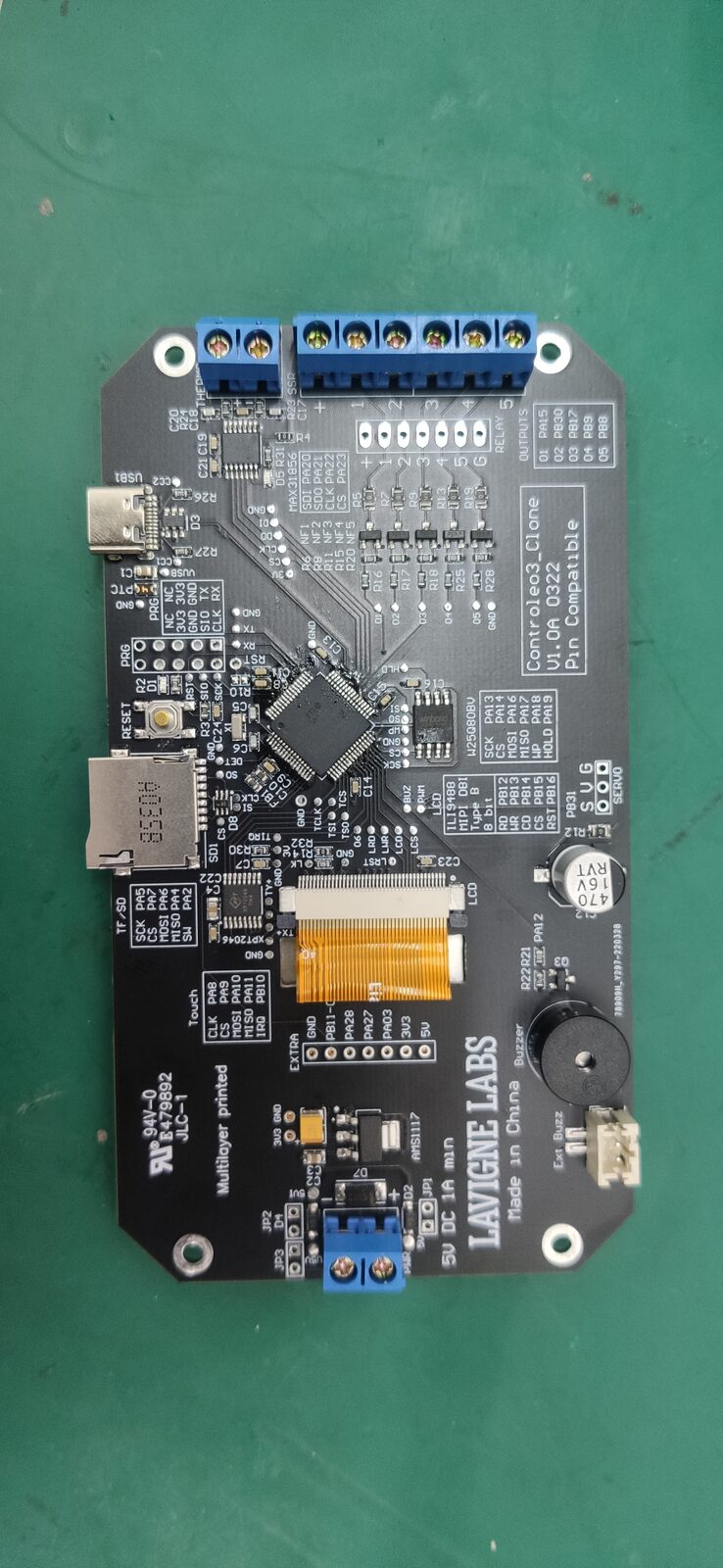 Black circuit board with screw terminals, USB-C port, a chip, an orange display ribbon and a buzzer on a green mat.