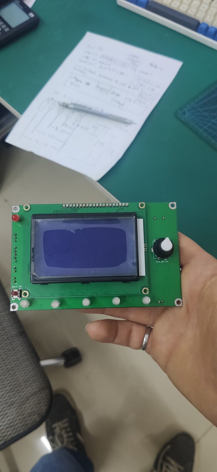 A hand holding a green circuit board with a blue character LCD, above handwritten notes.