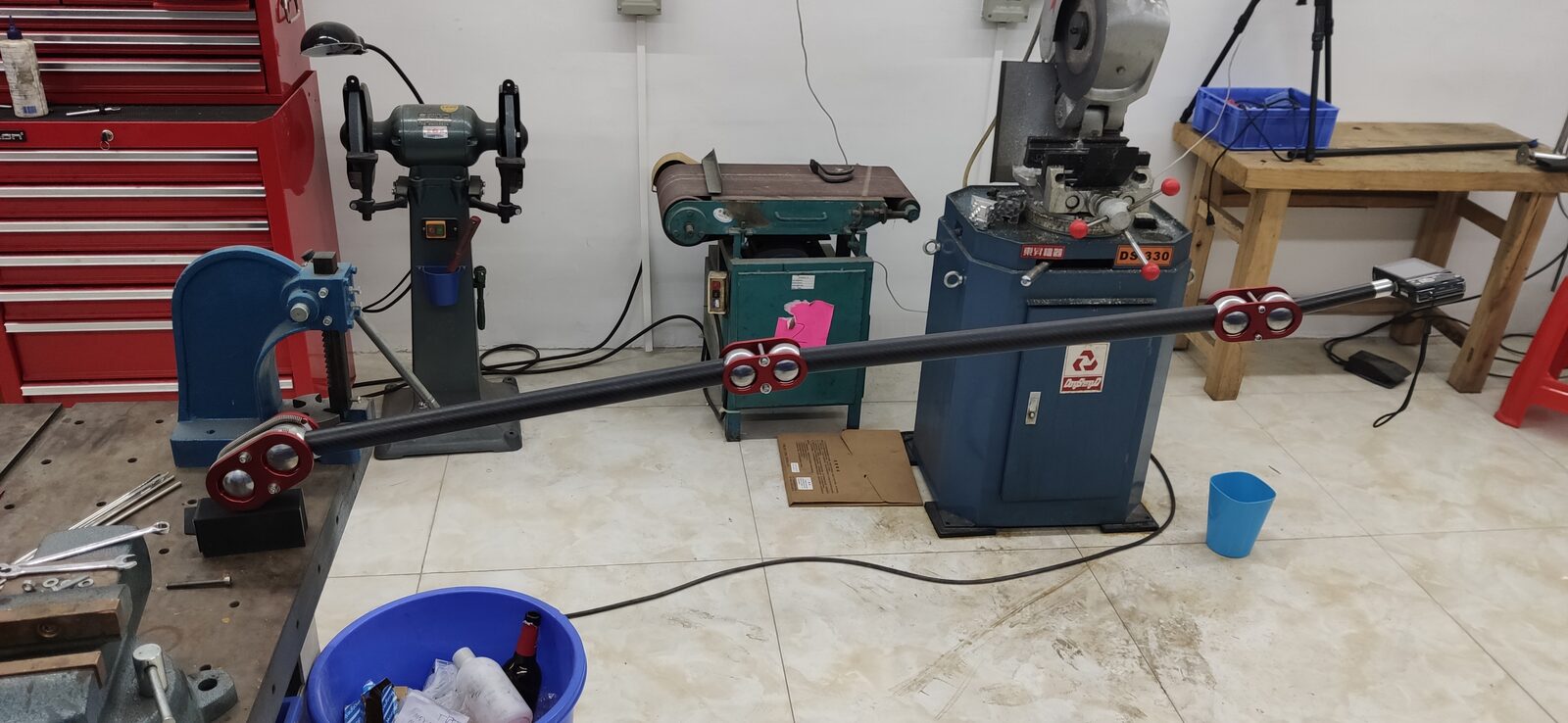 A second view of the long arm with red joints stretched across the workshop, with a blue bucket and machine tools nearby.
