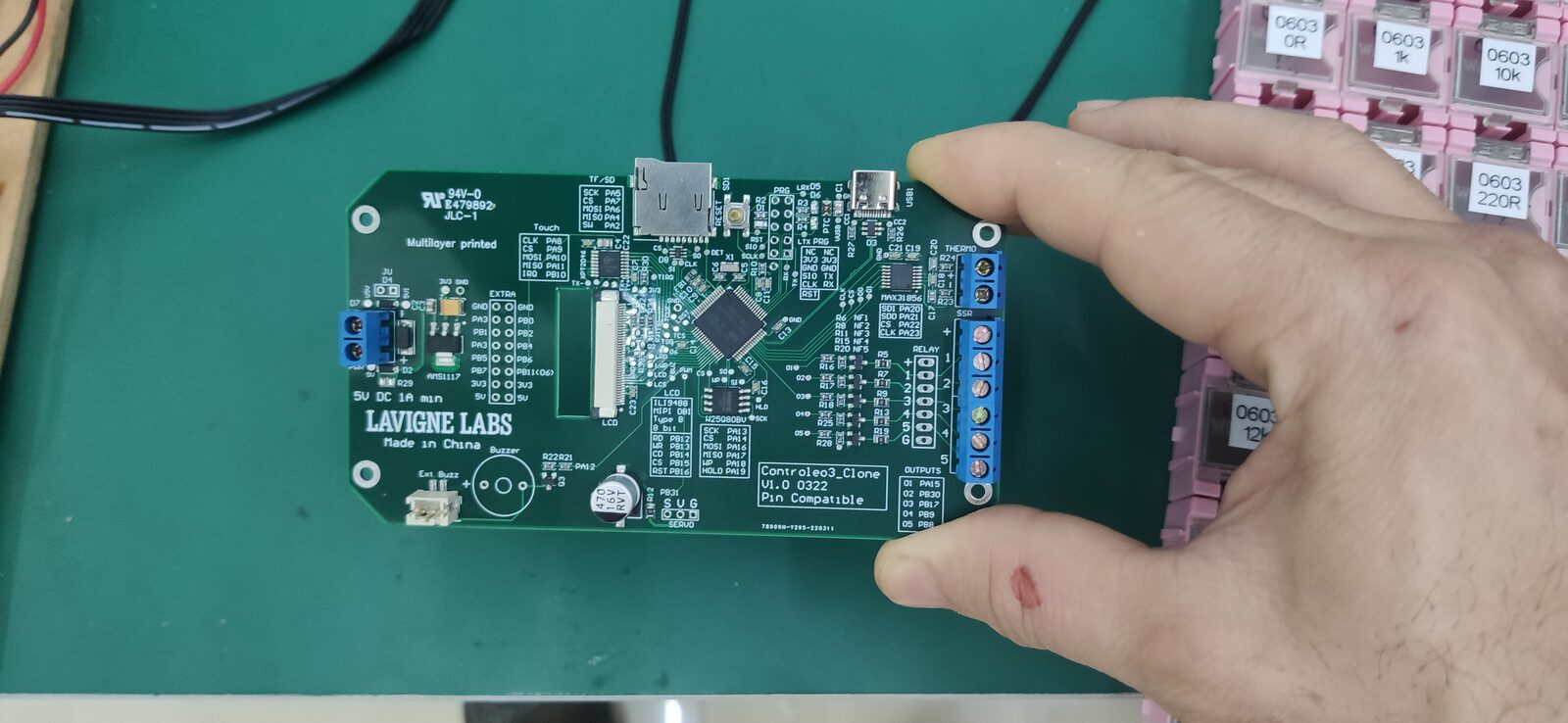 A hand holding a small circuit board with screw terminals, a connector and a black chip over a green cutting mat.