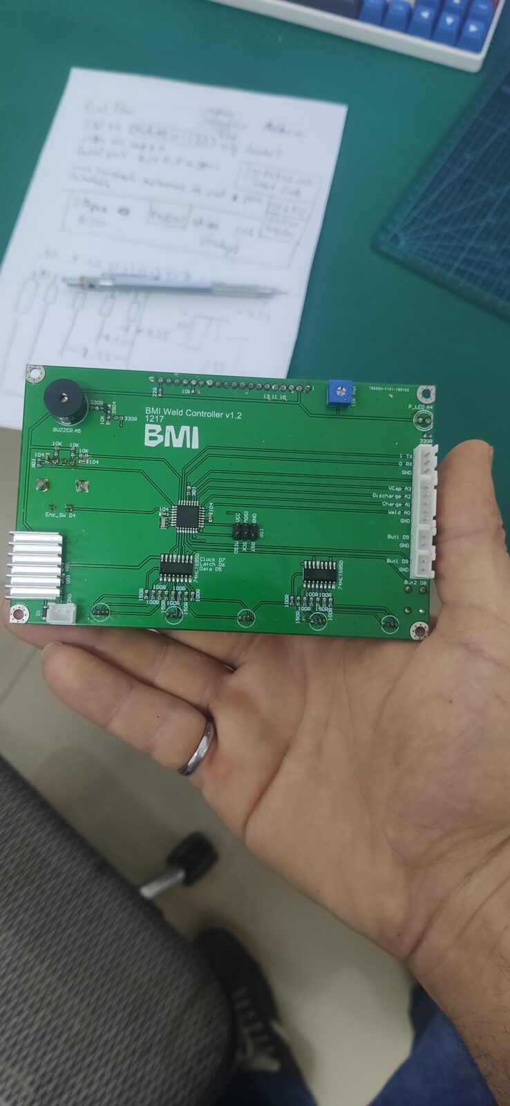 A hand holding a green circuit board with a row of header pins and pads, above a handwritten page on a desk.