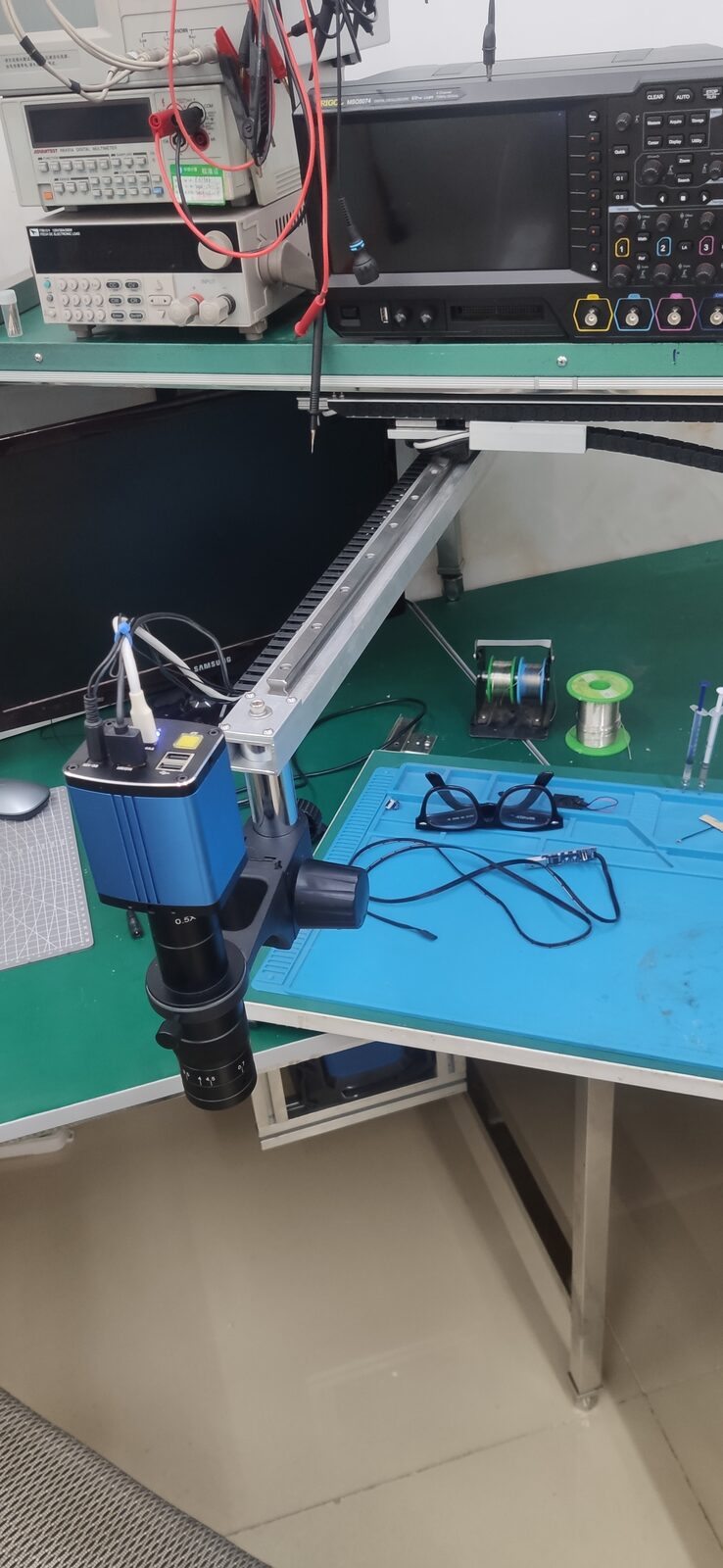 Microscope extended on a pale metal slide rail over the work mat, seen beside an oscilloscope and bench power supplies.