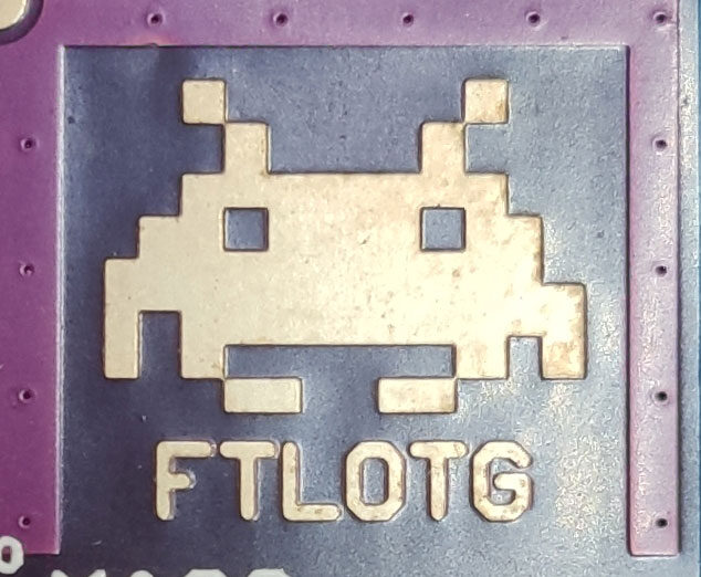 Close-up of a cream-coloured copper pixel-art space invader on a purple board, with FTLOTG below.
